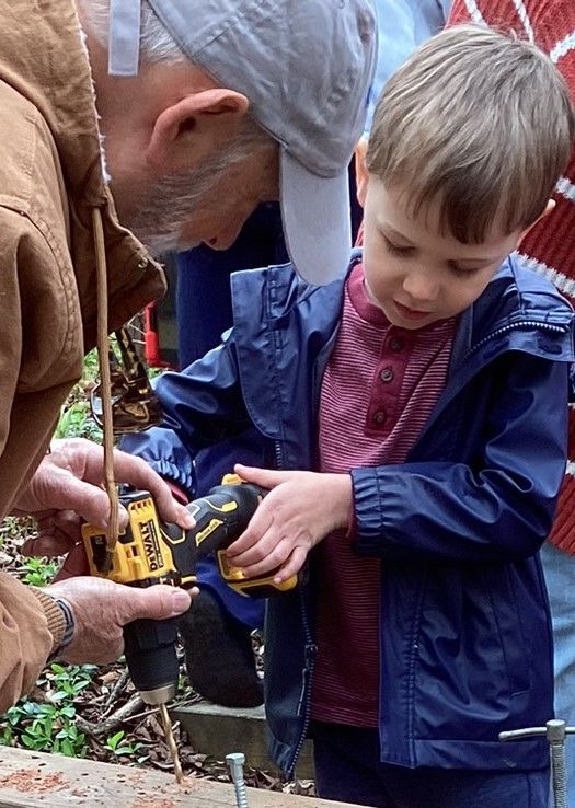 Young boy learning to use a drill with the help of an adult. Both are focused, with the boy wearing a blue jacket.