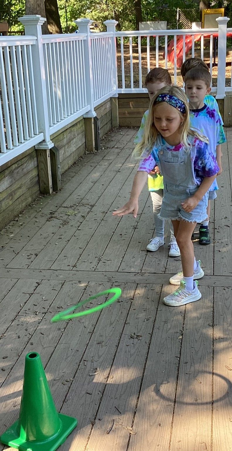 Children playing a ring toss game on a wooden deck. A girl throws a green ring towards a green cone, with two other children watching.