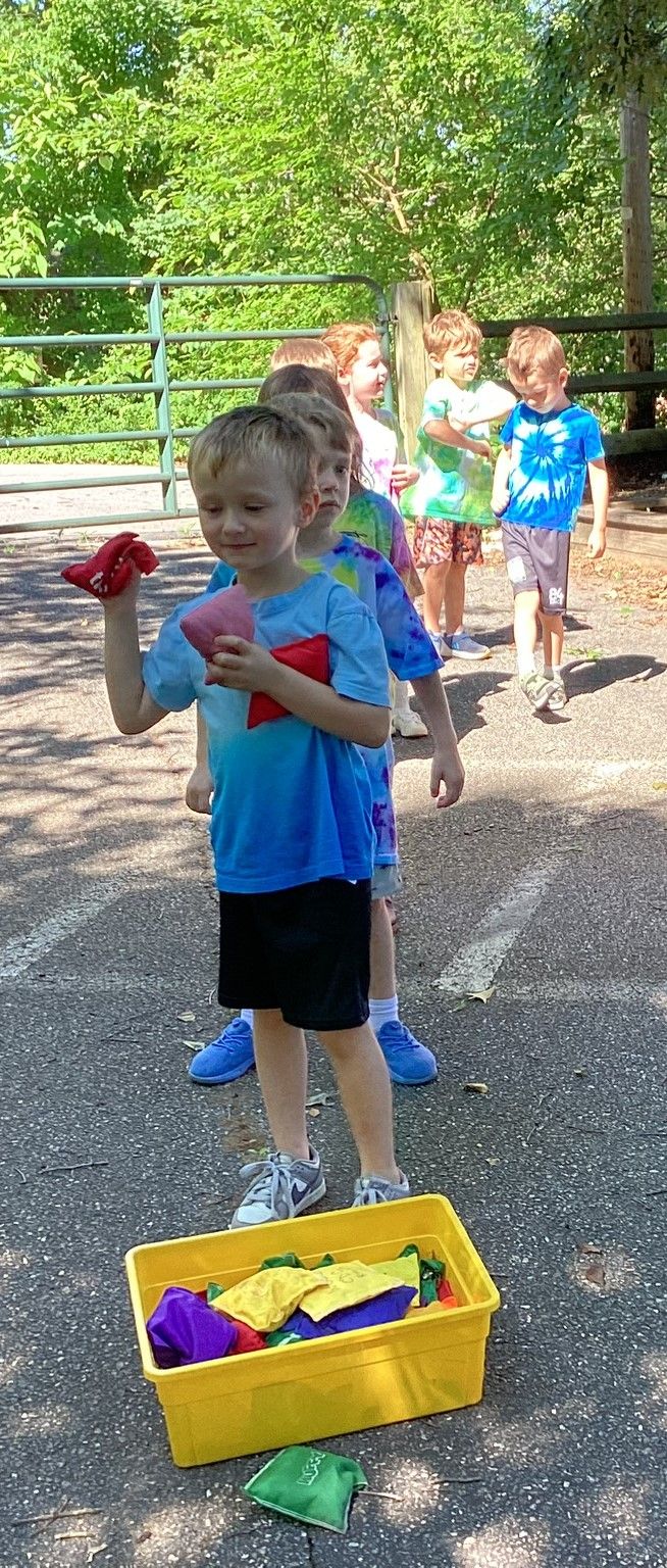 Children playing a bean bag toss game outdoors. A boy in a blue shirt holds a red bean bag, a yellow bin is on the ground.
