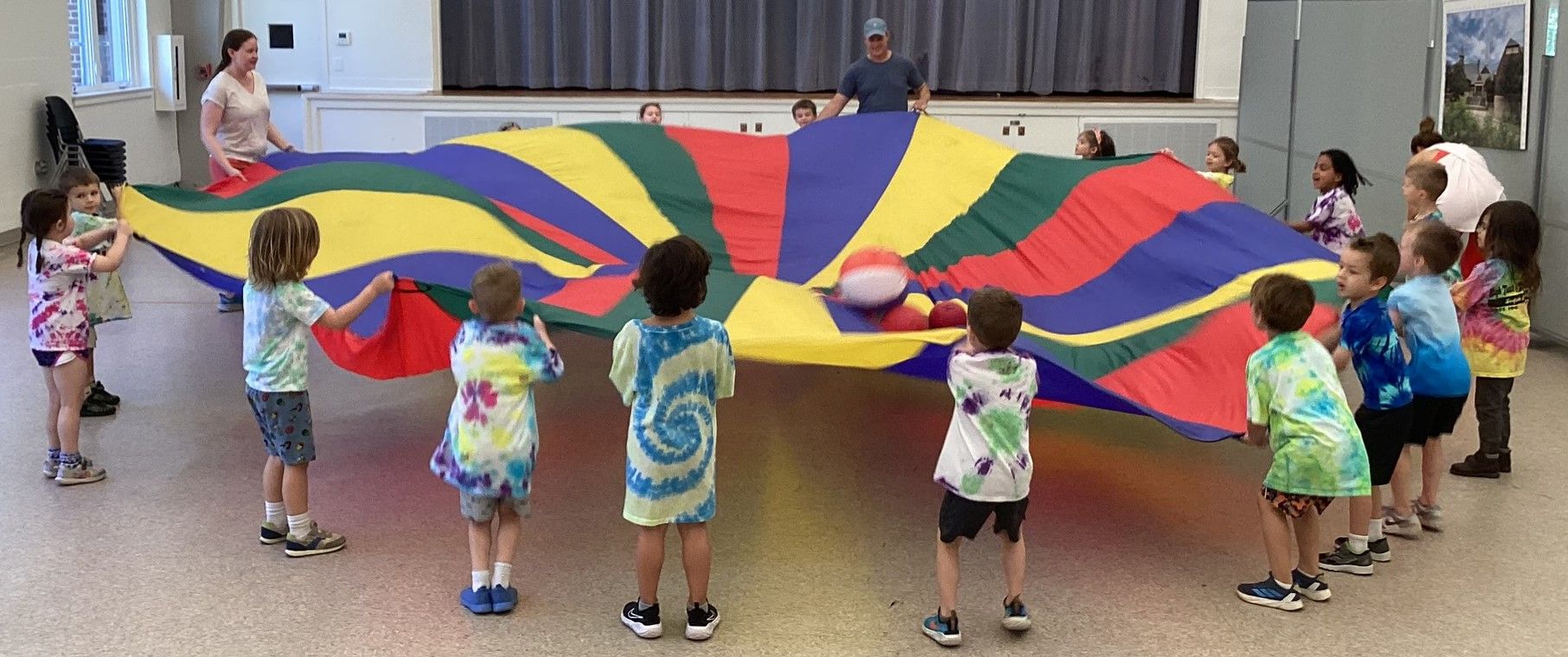 A group of children in tie-dye shirts play with a large, colorful parachute in a room with adults supervising.