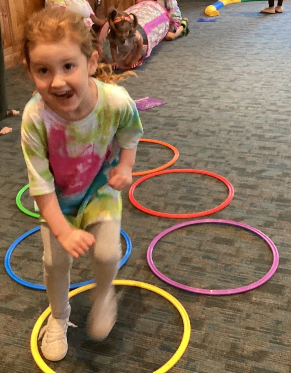 Young girl in tie-dye shirt happily jumps through colorful hula hoops on a carpeted floor.