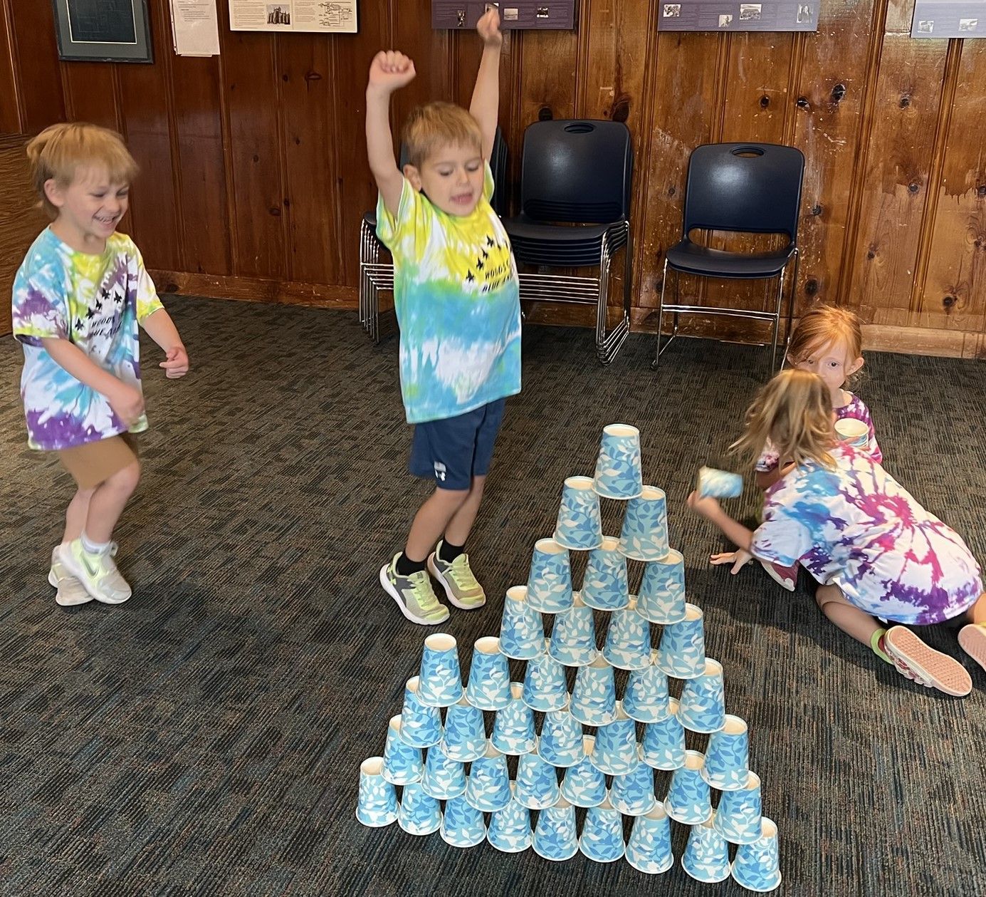 Children playing with a pyramid of cups. One boy cheers, others run or fall. They're in a room with wood paneling and chairs.