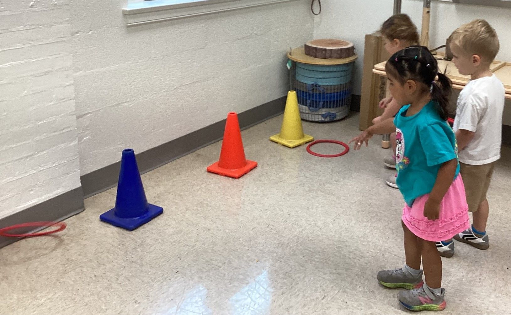 Children playing a ring toss game with cones in a classroom setting. A girl in pink skirt prepares to throw a ring.