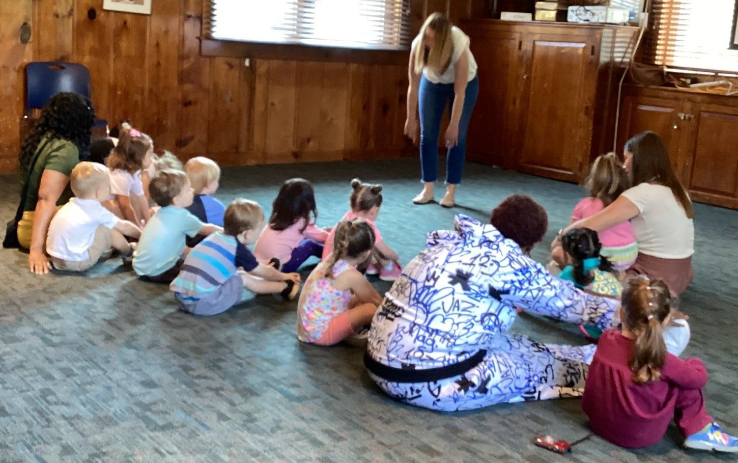 Children and teachers sit in a circle on a blue carpet in a room with wood paneling. A teacher is facing them.