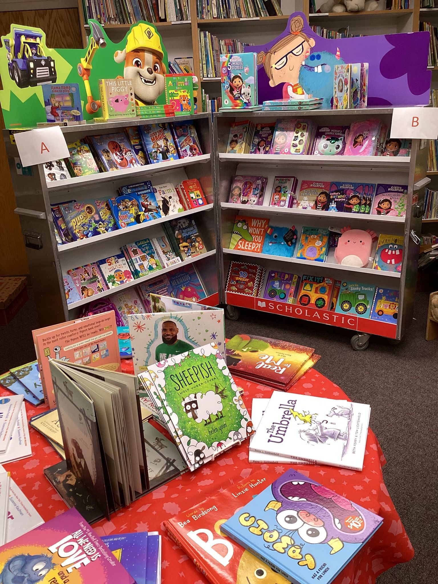 Children's books on display at a library book sale. Several shelves are filled with colorful book covers; additional books are on a red table.