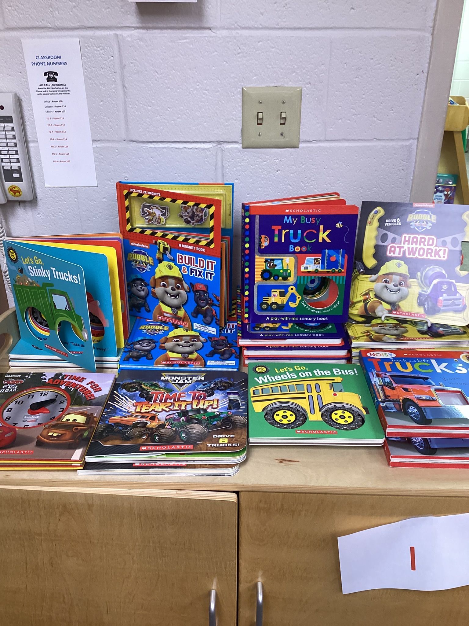 Books about vehicles and construction, featuring characters, on a wooden shelf against a white wall with an electrical outlet.
