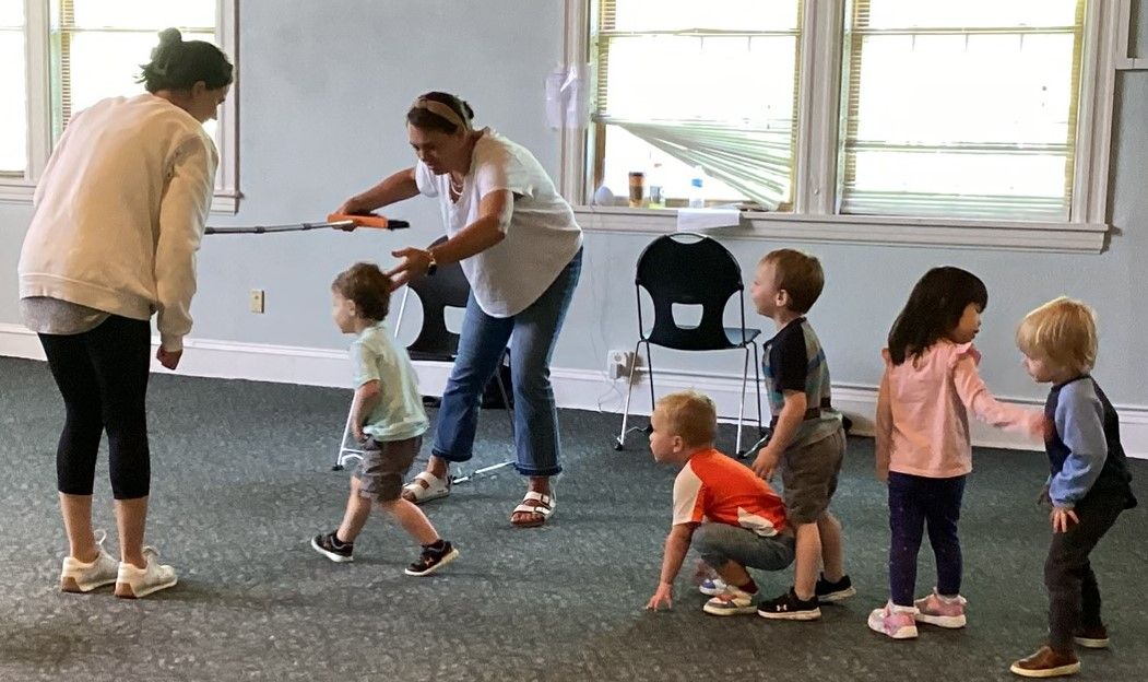 Two adults guiding a group of young children indoors; some are walking, crouching, or reaching towards others.