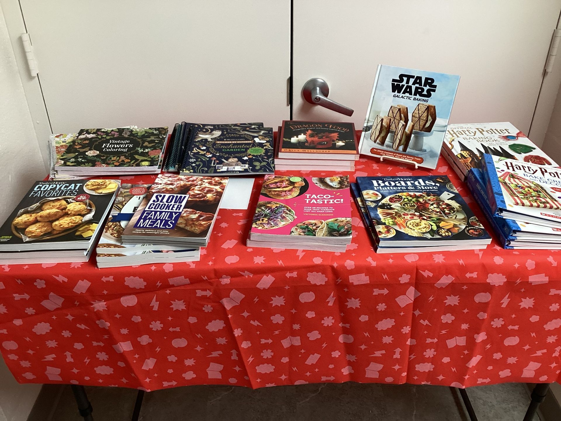 A table covered with a red cloth featuring several cookbooks, including a Star Wars-themed one, in front of a white door.