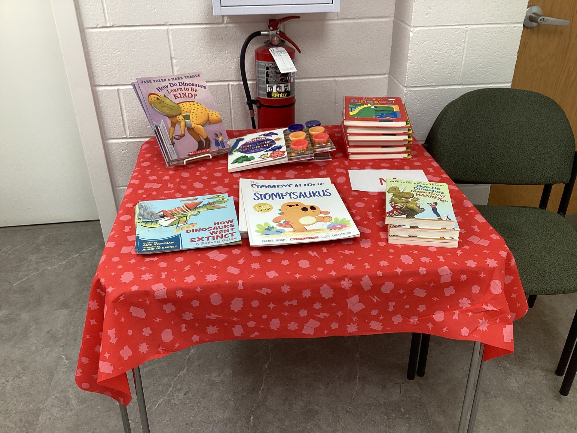 A table covered in a red cloth displaying children's books, with a fire extinguisher in the background. A green chair sits next to the table.