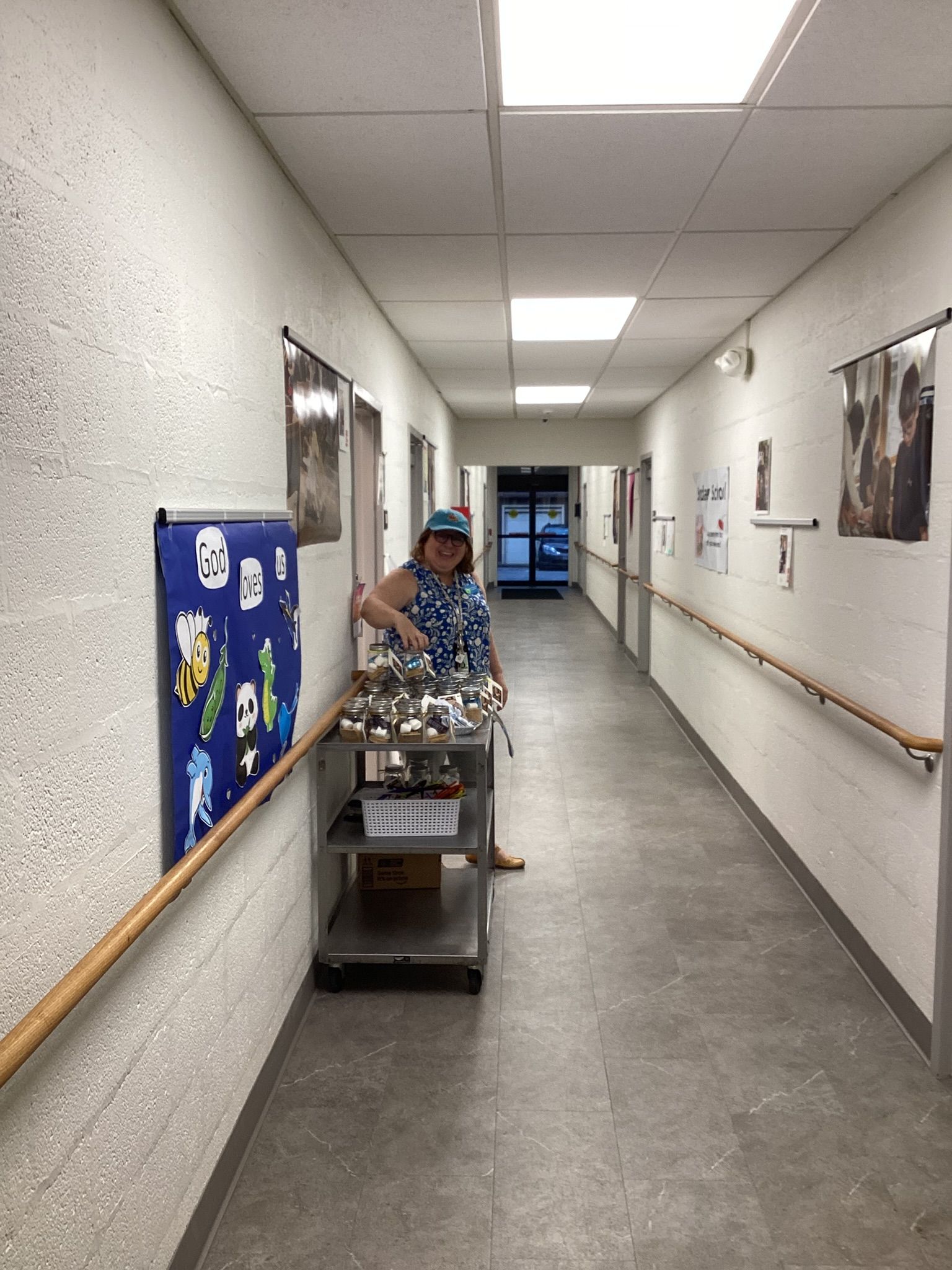 Person with a food cart in a hallway. They are smiling while offering food from the cart. The walls are light and there are framed pictures hanging.