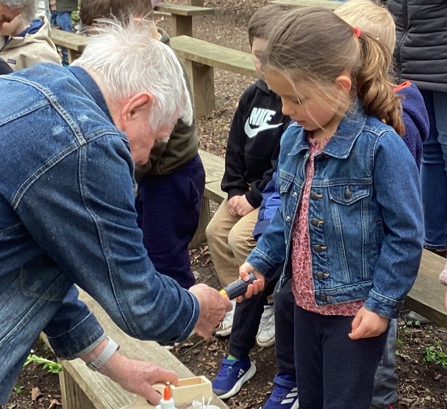 An older man assists a young girl in an outdoor setting. Both are looking down at a small object. Other children are in the background.