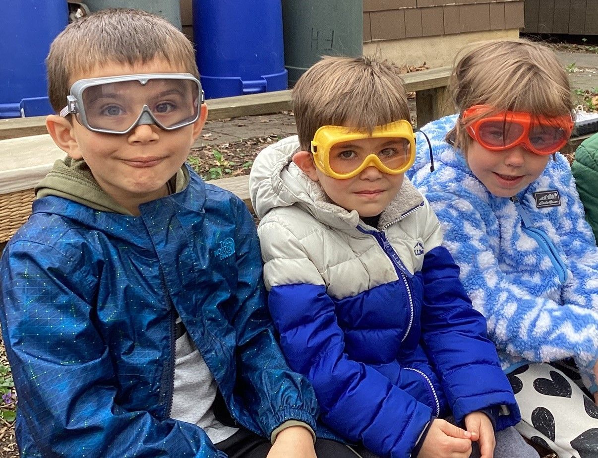 Three young children wearing safety goggles, outside. They're bundled in jackets, smiling, and sitting on a bench.