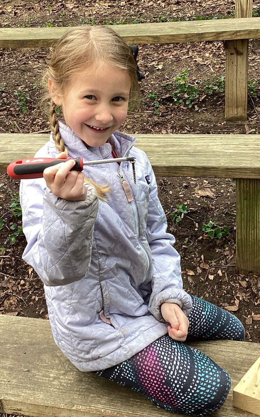 Young girl with blonde braids smiles, holding a screwdriver while sitting on a wooden bench in a wooded area.