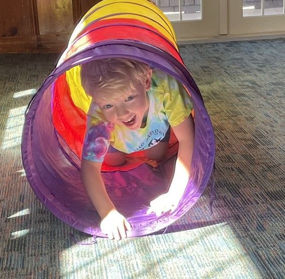 Young child smiling, crawling through a colorful play tunnel on a carpeted floor.