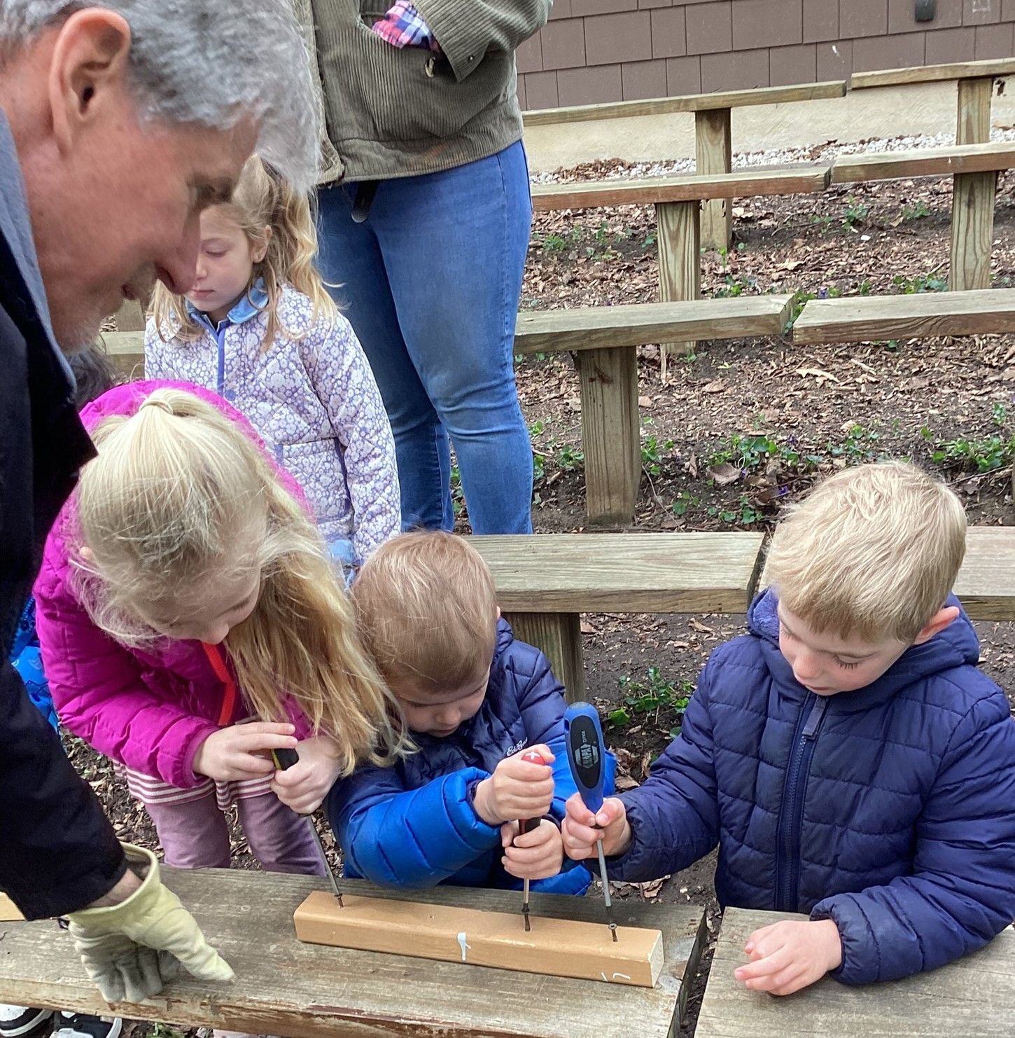 Children and an adult work together, hammering nails into a wooden plank outdoors. Several other children observe.