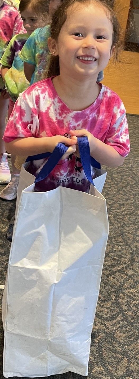 Young girl smiling, wearing a pink tie-dye shirt, holding a white bag with blue handles.