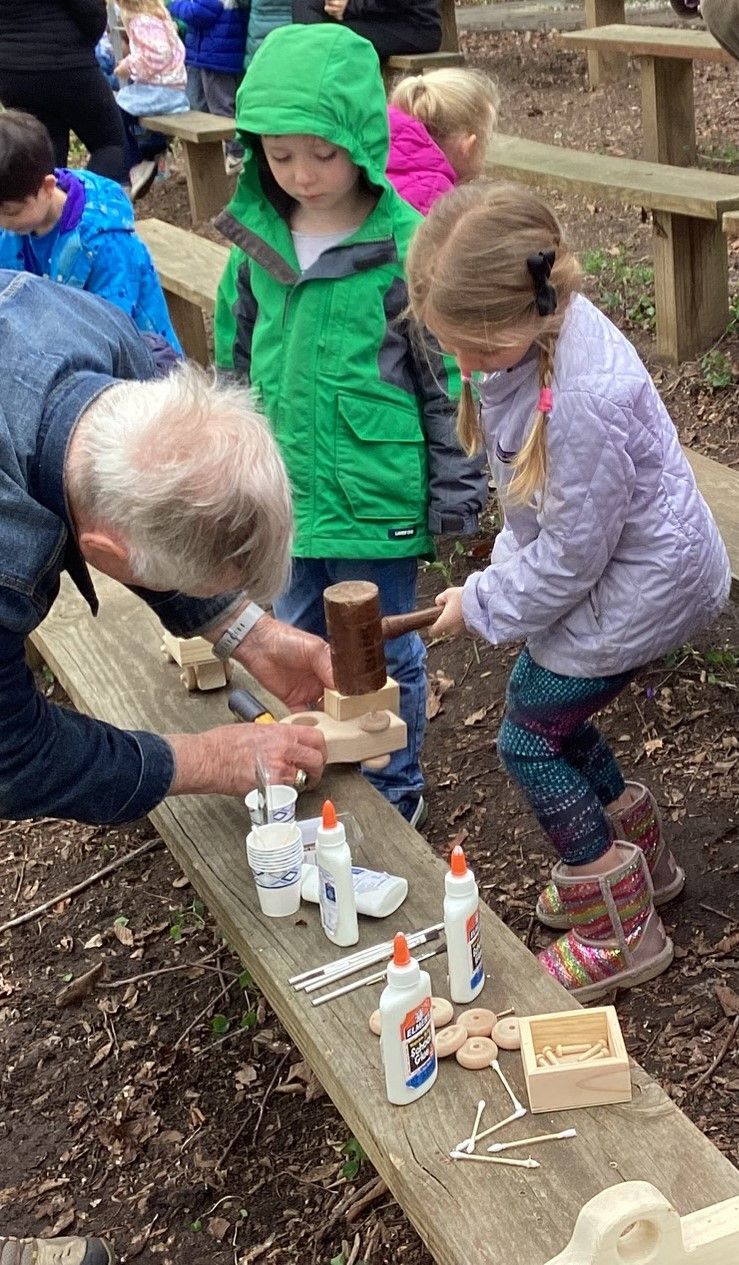 A man helps a girl hammer wood pieces on a bench. Other children watch, some wearing colorful jackets.