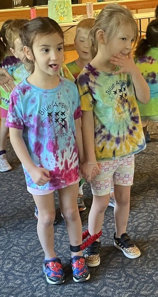 Two young girls in tie-dye shirts and shorts, holding hands. They stand inside, possibly at a party, with others visible in the background.