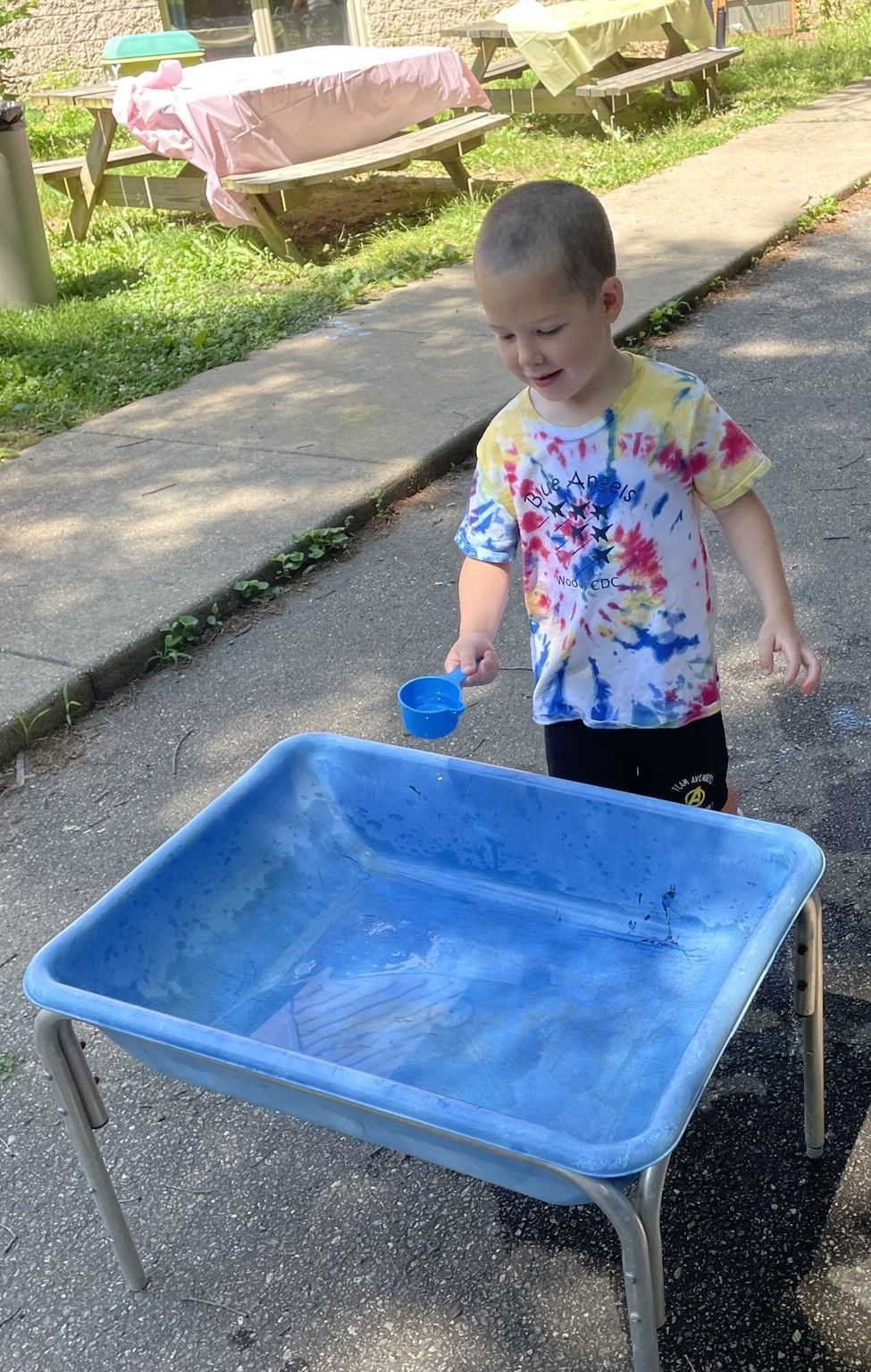 A young boy in a tie-dye shirt smiles while playing with water in a blue tub outdoors.