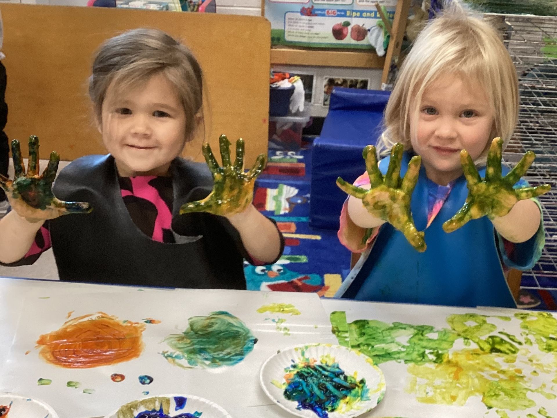 Two young children with paint-covered hands smiling, working on a craft at a table. Various colored paints visible.