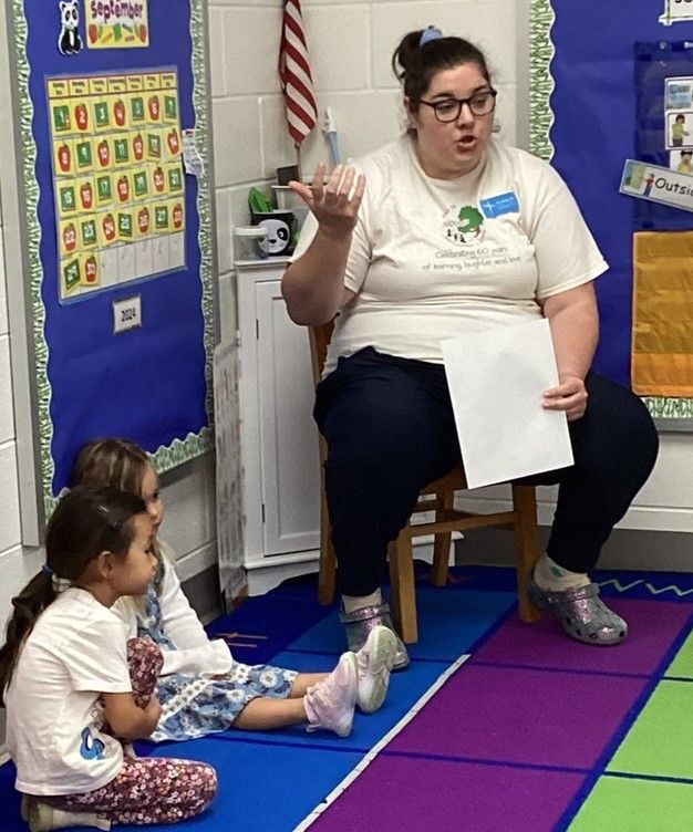 A teacher in a classroom points while reading to two young students seated on the floor.