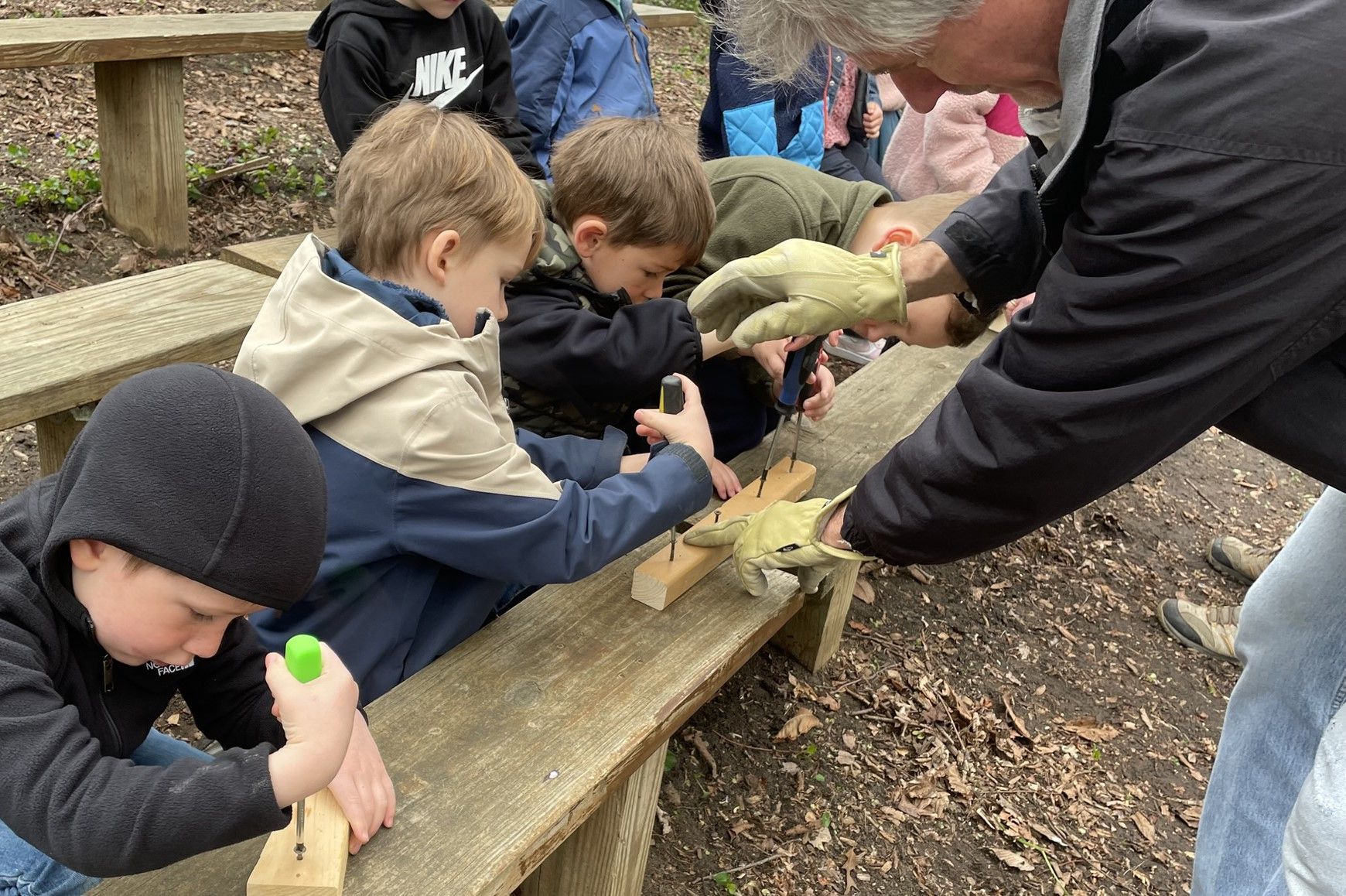 Children and an adult are working together on a wooden project outdoors. The kids are using tools like small saws and looking intently.