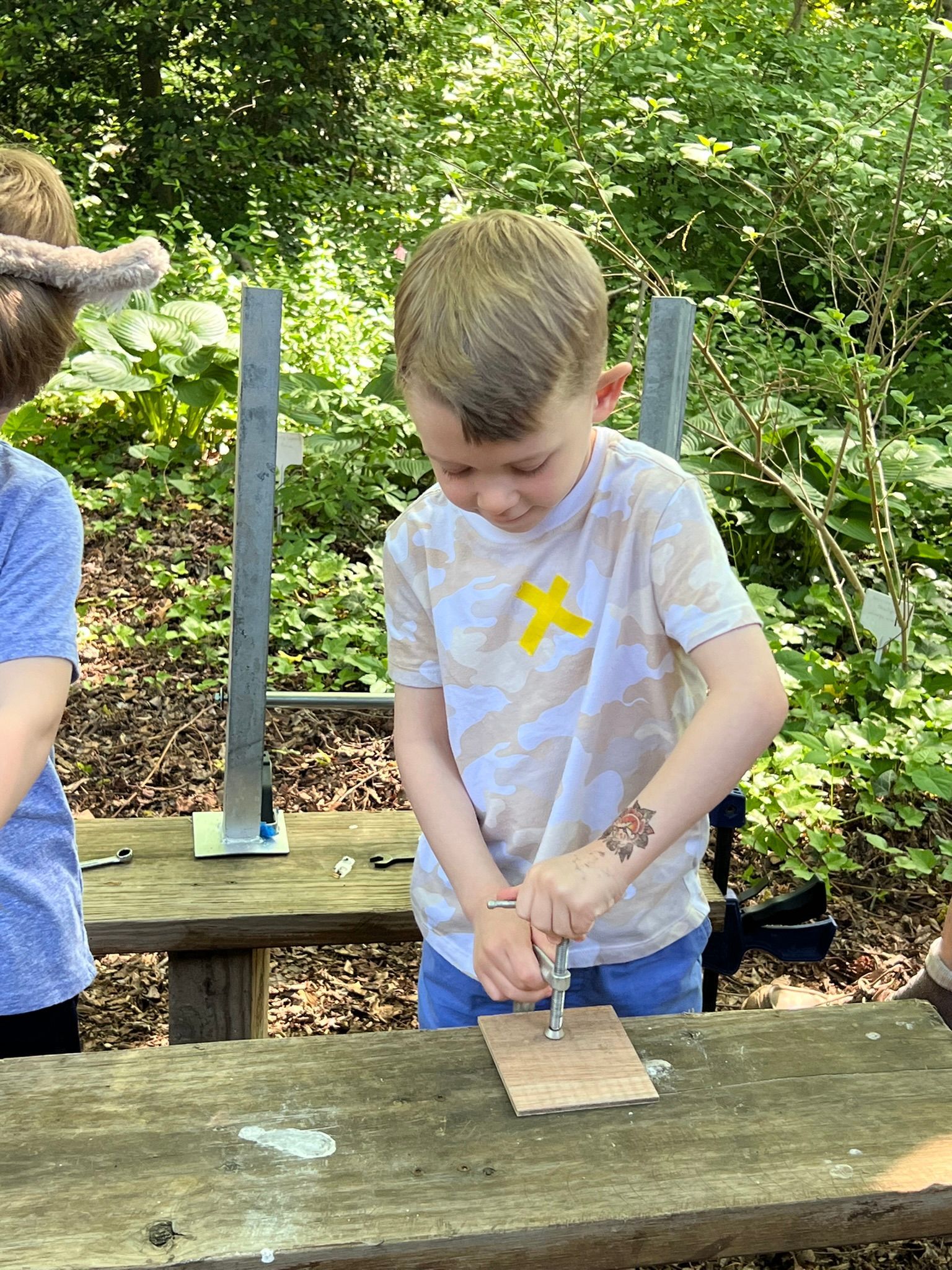 A young boy with blonde hair, wearing a light-colored shirt, screws something into a piece of wood on a wooden table outside.