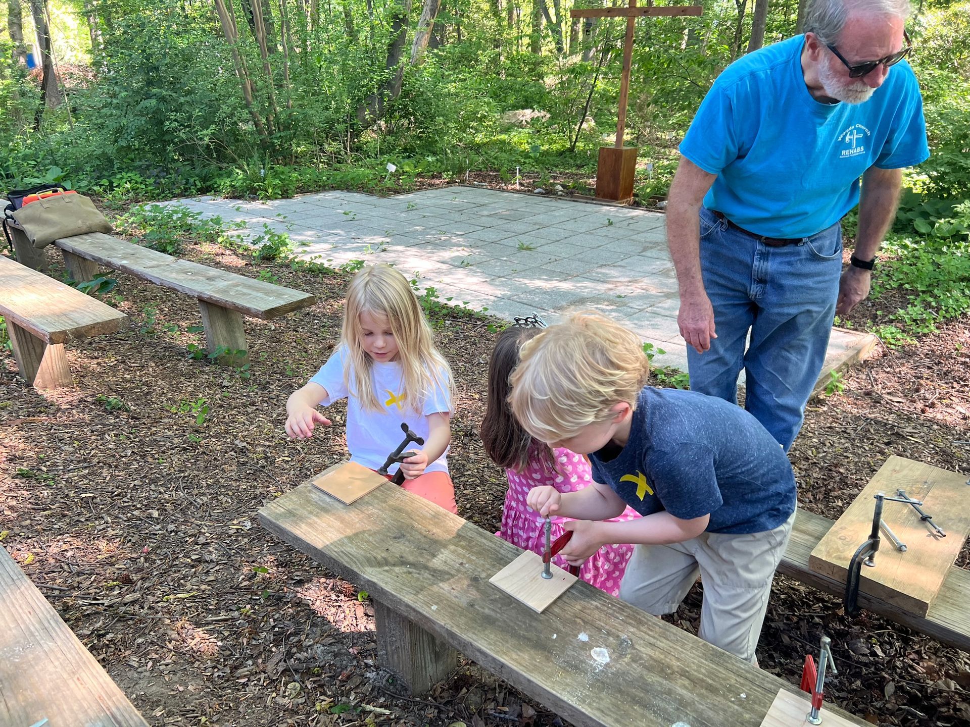 Three children and a man build with wood outdoors. The children, with tools, are on a wooden bench as the man watches.