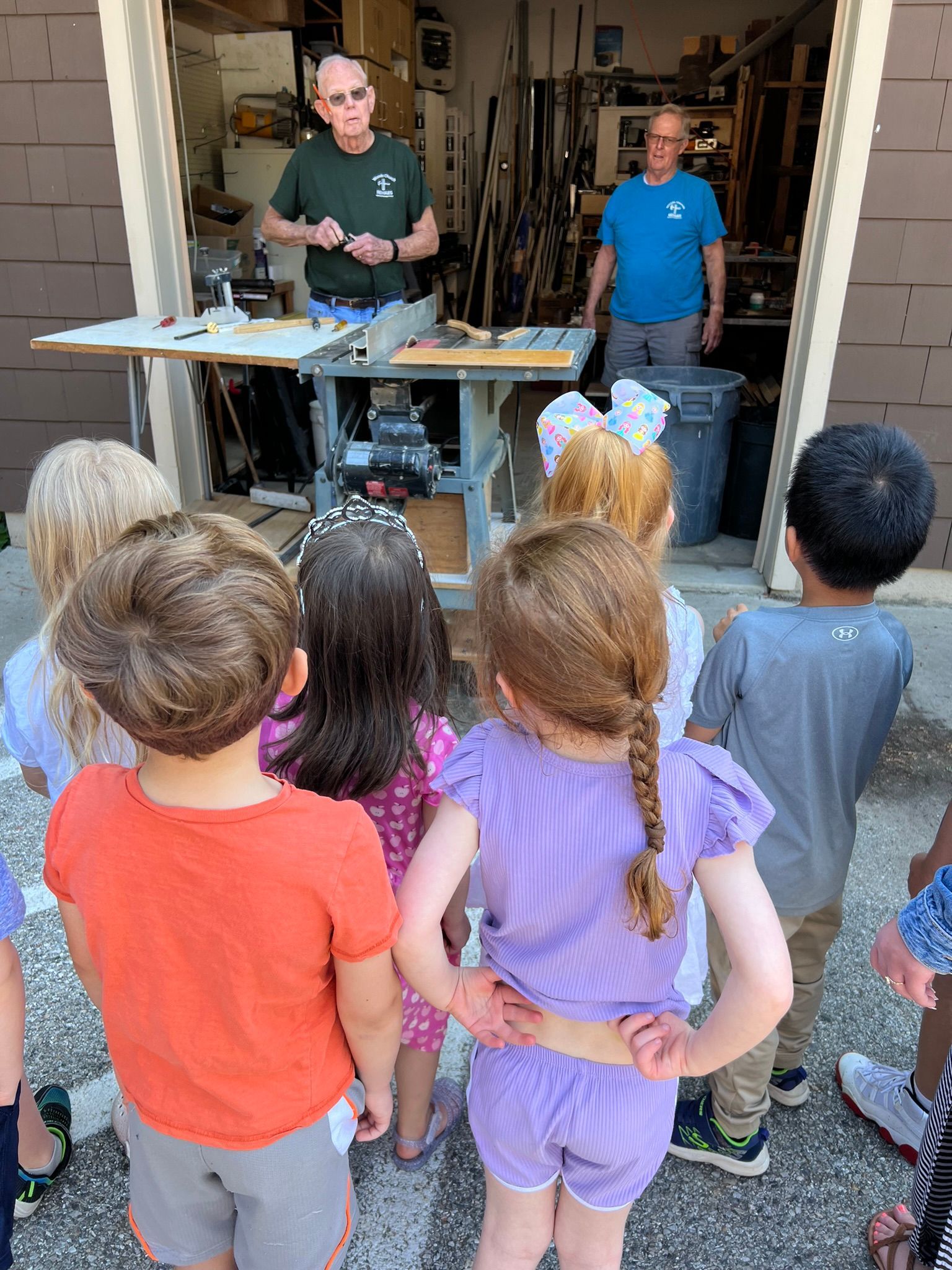 Children watch two men demonstrate woodworking in a garage. The men stand behind a table with tools, while the kids face them, some with pigtails and bows.