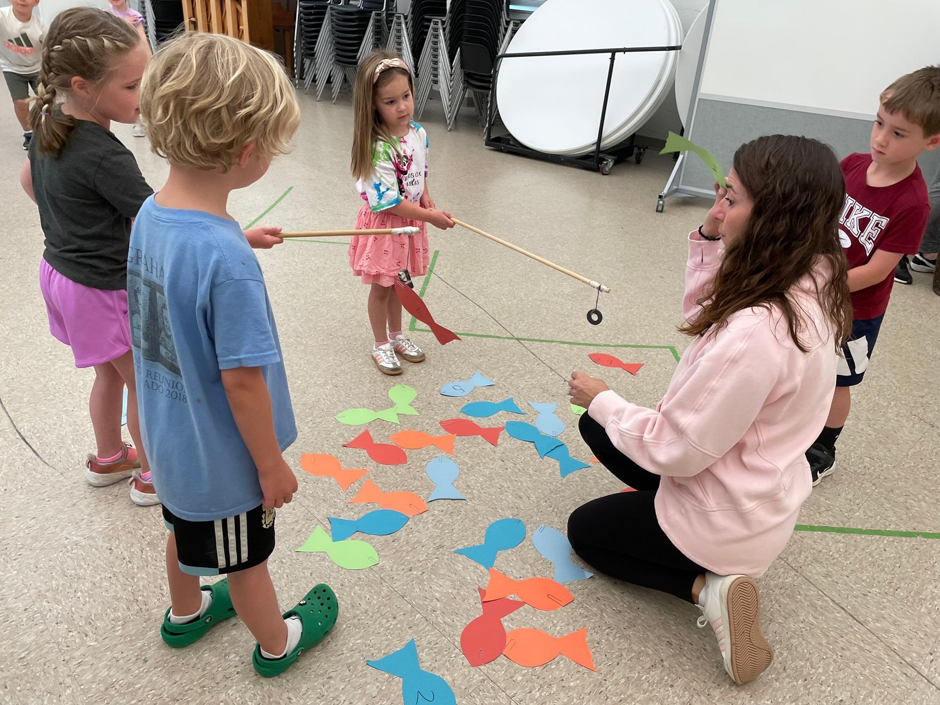 Children playing a fishing game with a teacher indoors. They are using a fishing rod to catch colorful paper fish on the floor.