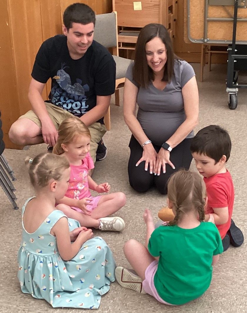 A man and pregnant woman sit with a circle of children on a floor. The adults smile and interact with the kids.