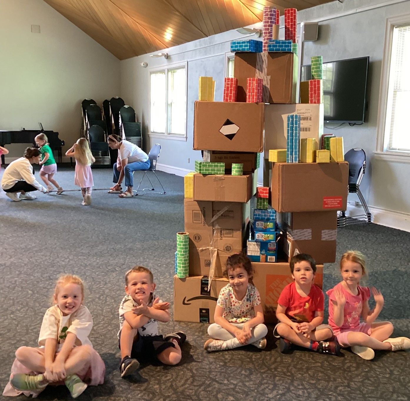 Children sit in front of a tower made of boxes and paper rolls. Other children and adults play in the background of a room with windows.