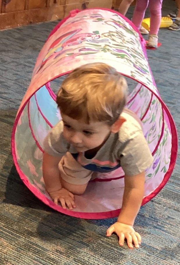 A young child crawls through a pink and patterned play tunnel on a blue carpet.