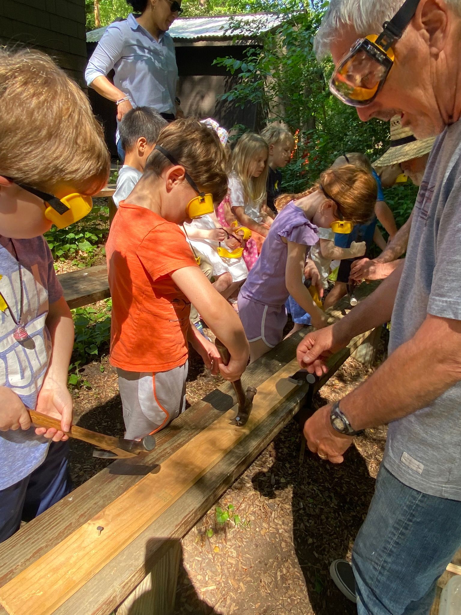 Children and an adult wearing goggles carve wood outdoors, guided by another adult. Some children wear safety glasses.