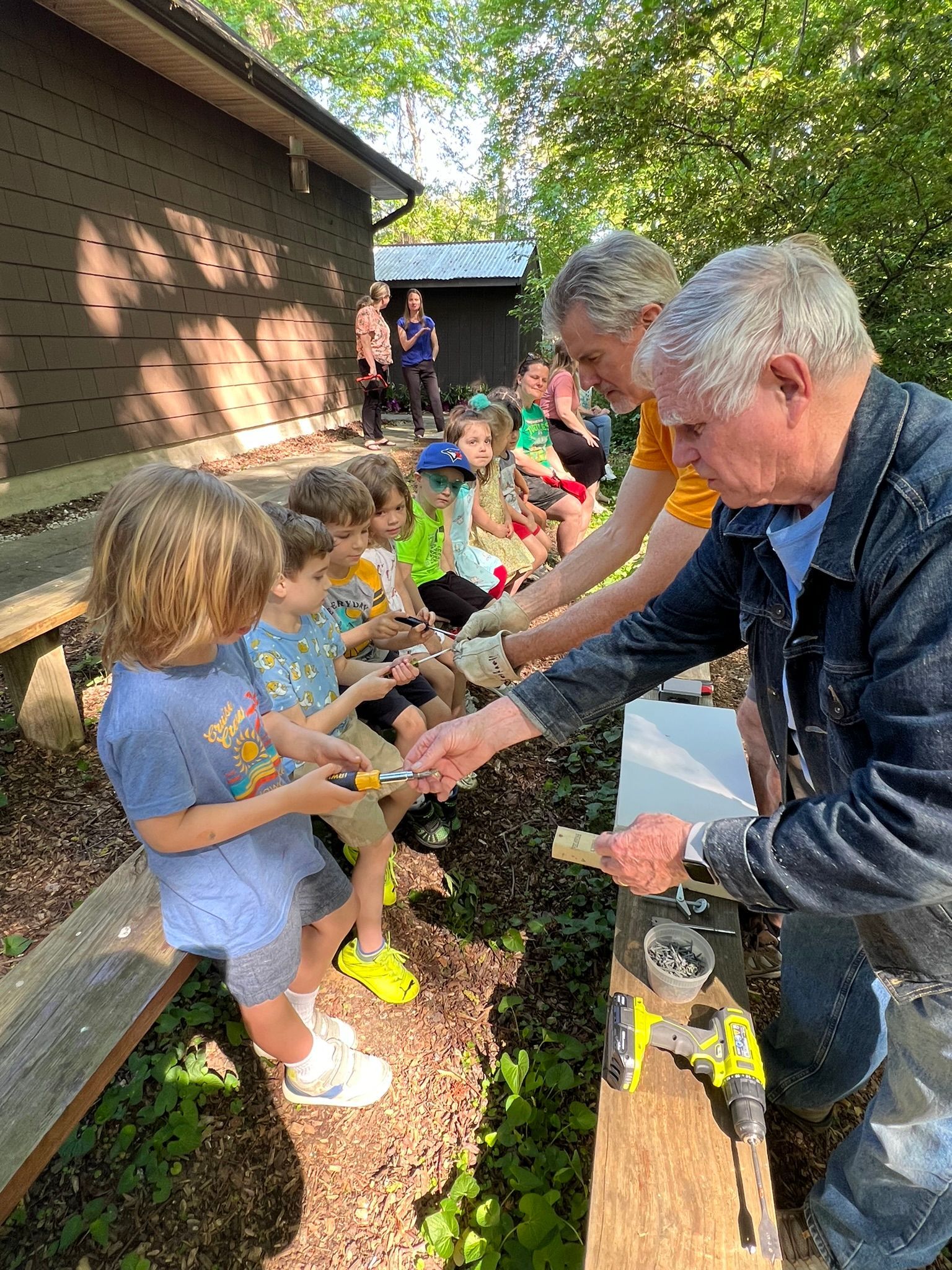 A group of children and adults outside, sharing tools and working on a project. Some kids are holding tools and a few adults are helping.