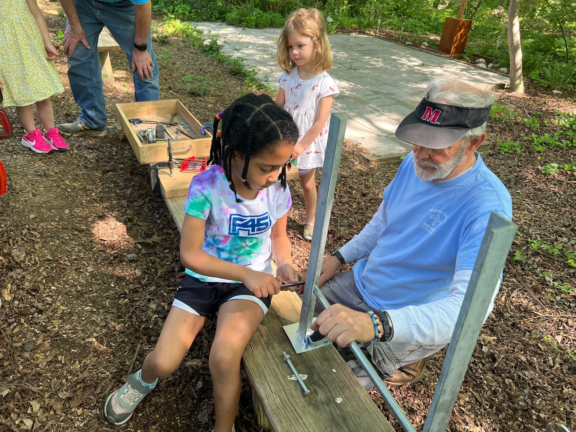 Girl and older man working on wood project outdoors. Another girl watches.