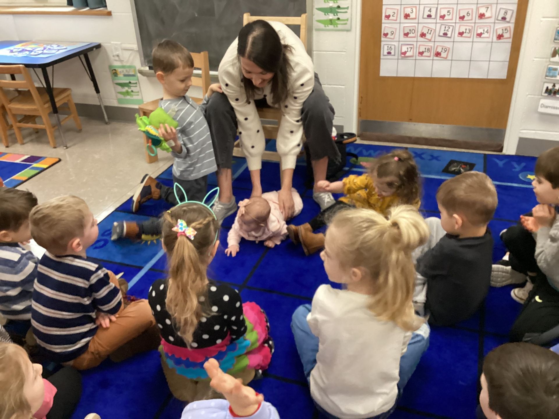 A teacher interacts with a baby on a blue mat surrounded by several children in a classroom setting.