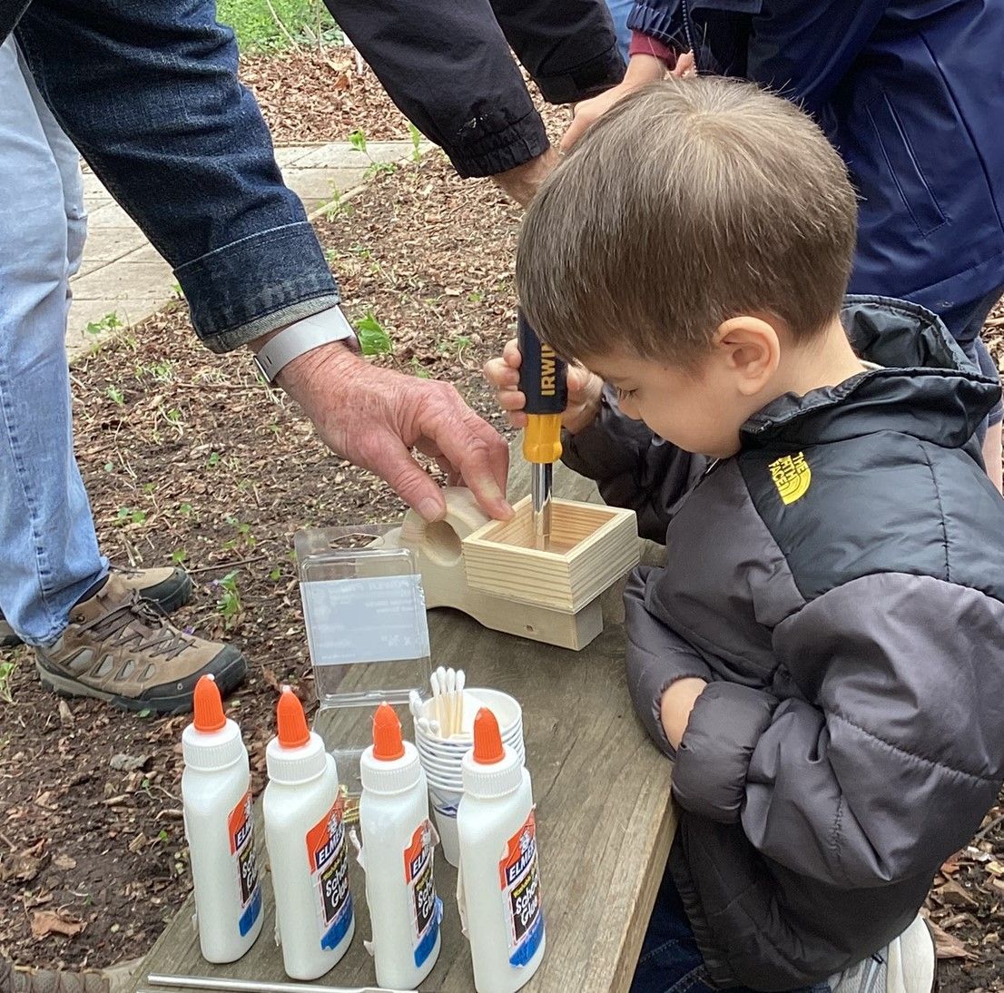 A young boy uses a screwdriver on a small wooden structure with help from an adult, surrounded by glue bottles and craft supplies.