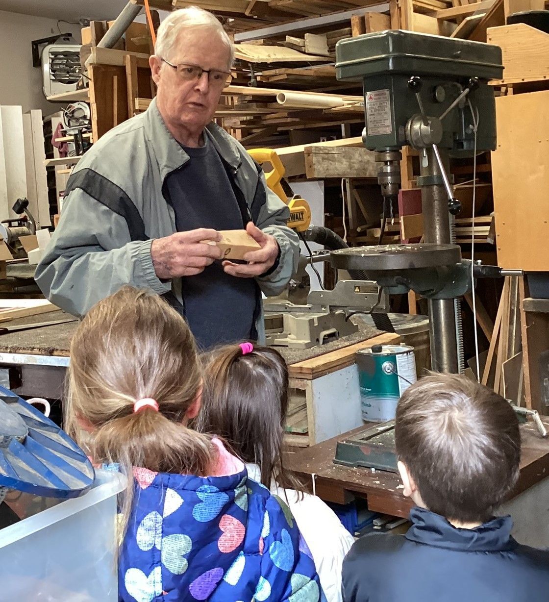 A man demonstrates woodworking to a group of children near a drill press. He holds a small wooden block while explaining.