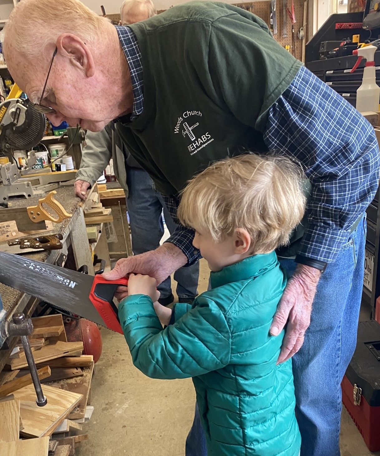 An elderly man helps a young child use a hand saw in a workshop. The man is wearing an apron and the child a green jacket.