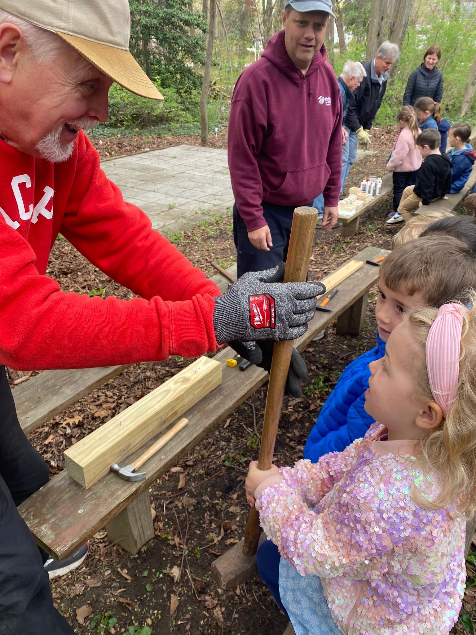 Man in red sweater demonstrates using a wood tool to a young girl, with other children and adults watching in an outdoor setting.