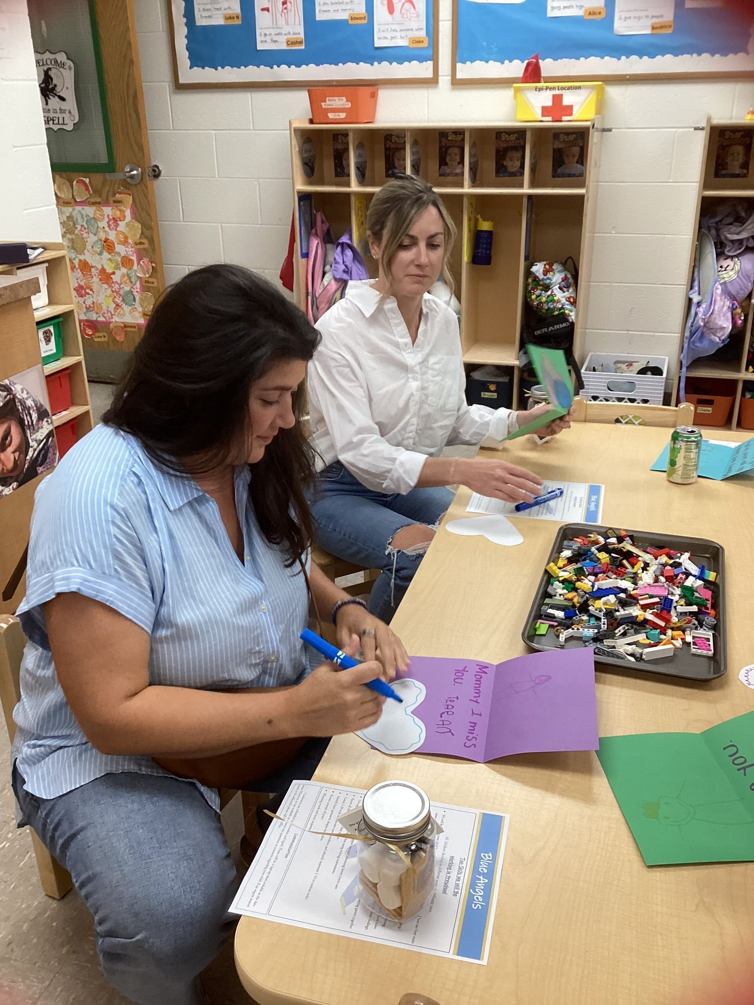 Two women seated at a table in a classroom, crafting. One writes on purple cardstock, the other sorts through green paper.