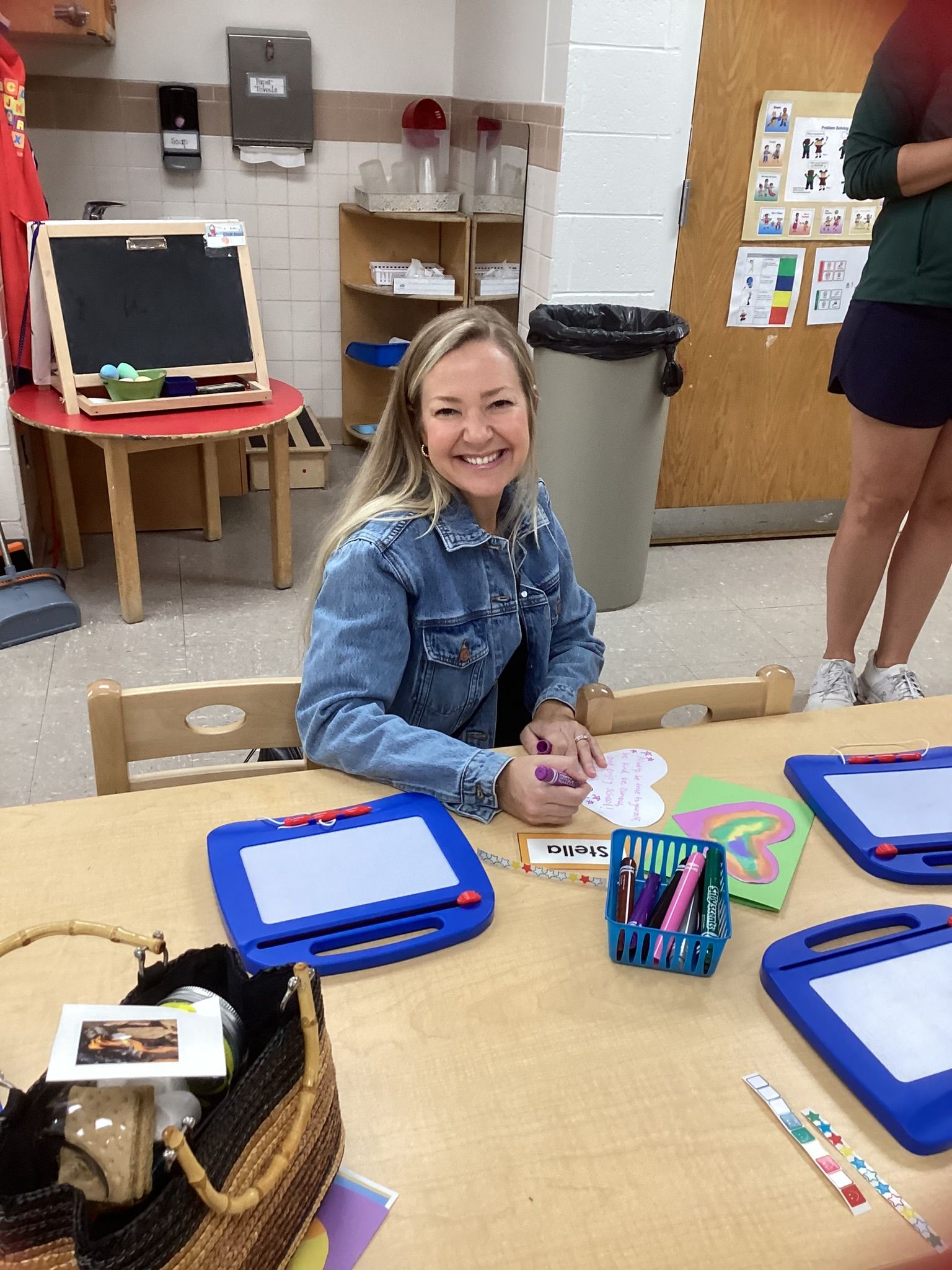 Woman in denim jacket smiling, drawing at a table with art supplies in a classroom setting.