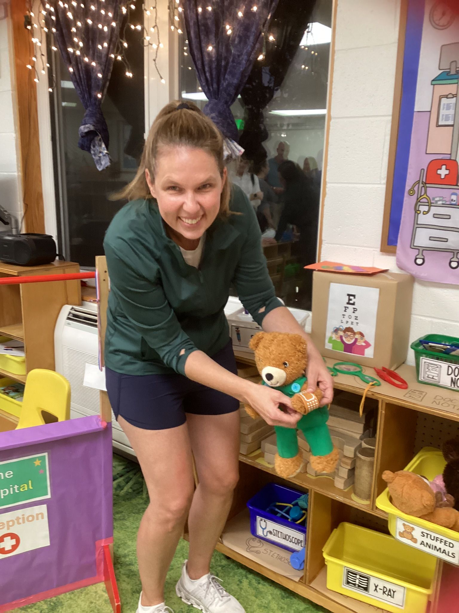 Woman in green shirt and shorts holds a teddy bear in a brightly lit classroom. She smiles as if tending to the bear.