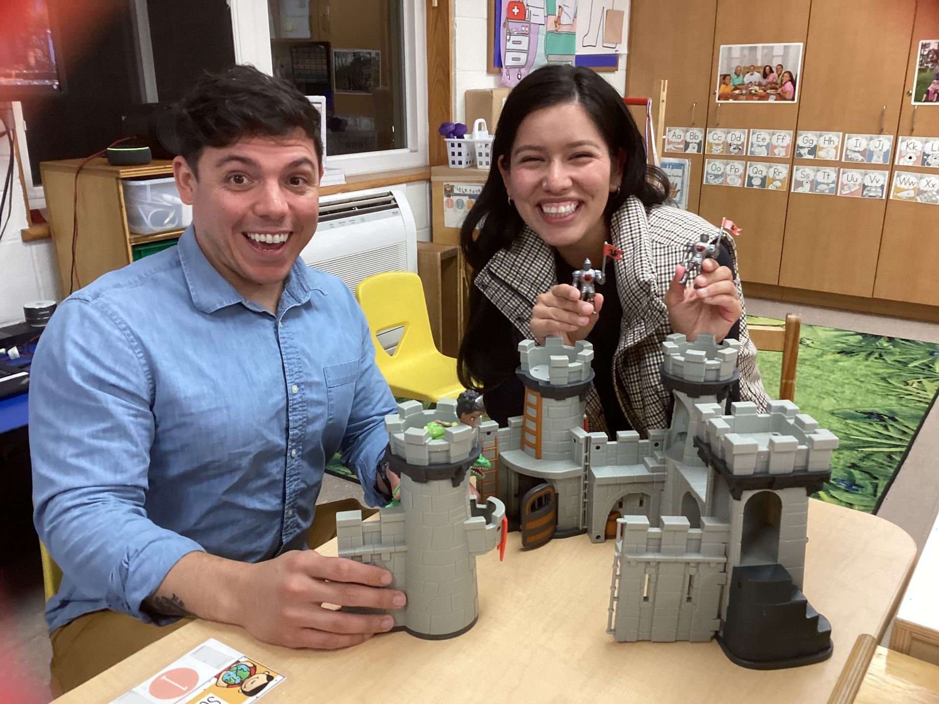 Two smiling adults build a toy castle in a brightly lit room. The man wears a blue shirt, the woman a patterned top.