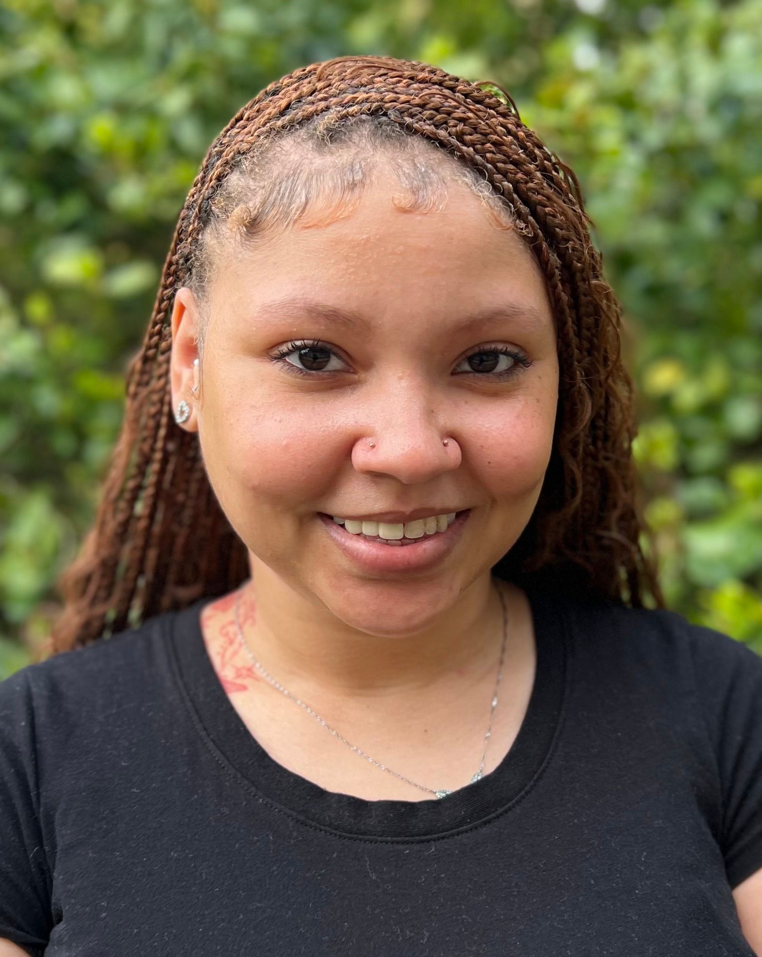 Black woman with long braided hair, nose piercings, and a blue lanyard smiles at the camera in front of leafy green foliage.