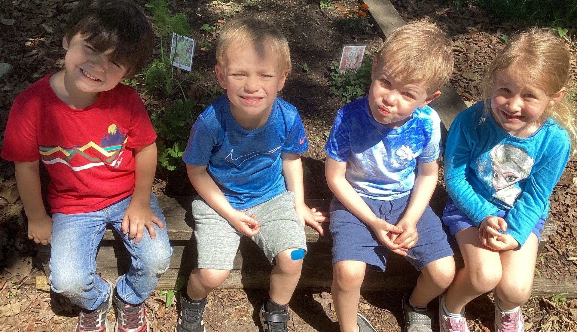 Four young children sitting outside on a wooden bench, smiling at the camera. They are wearing colorful shirts and are in a garden setting.
