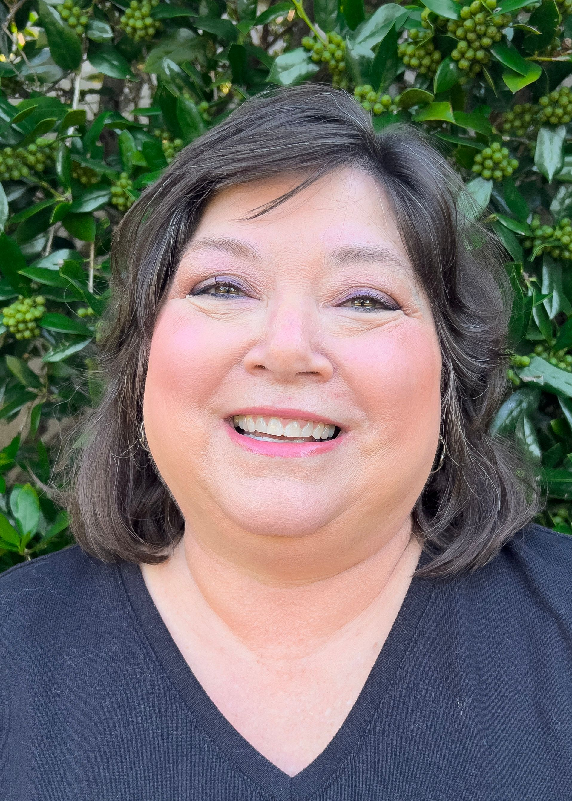 Woman with dark hair smiles at the camera against a green leafy background.
