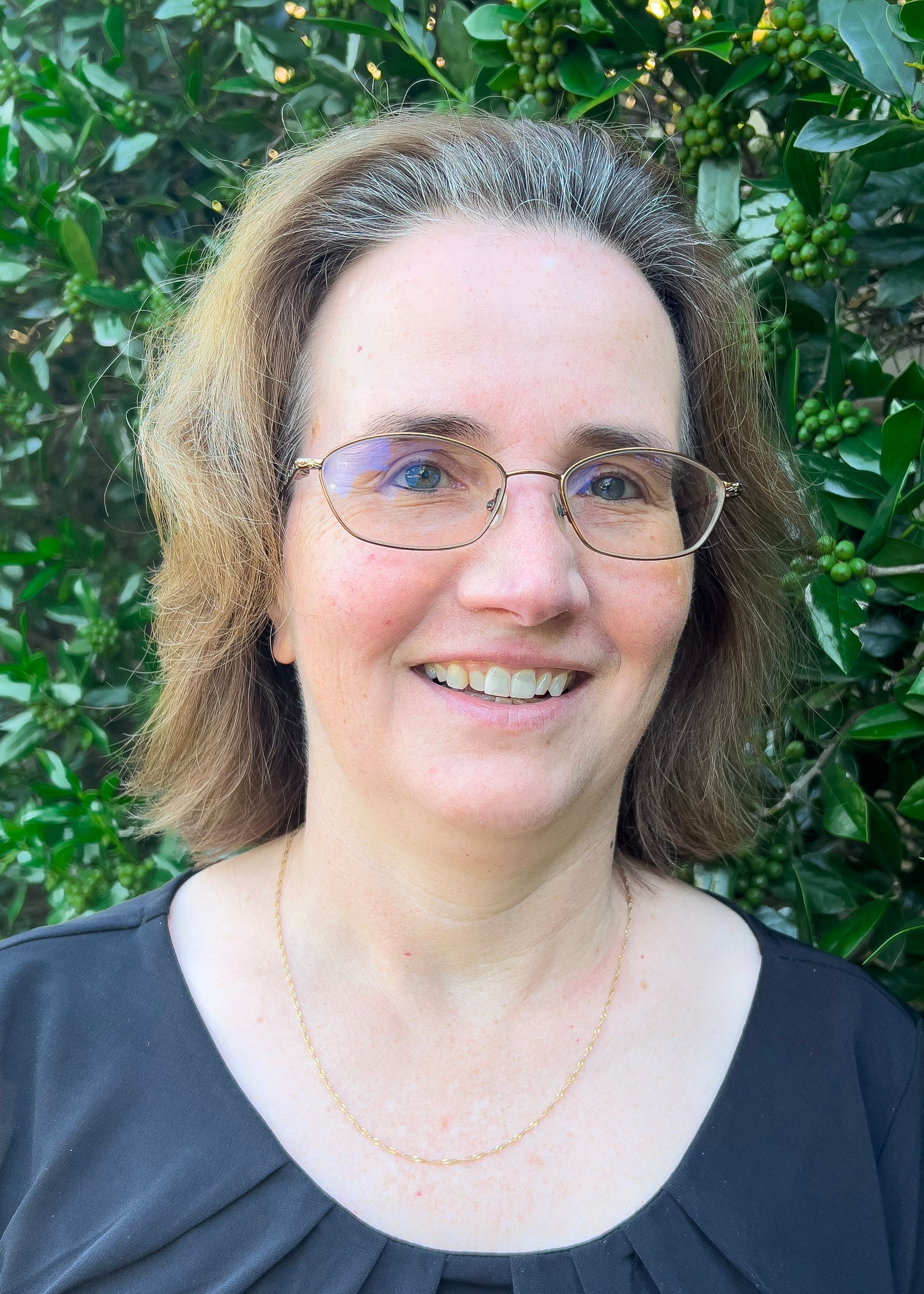 Woman with glasses and short brown hair, wearing a camouflage t-shirt, smiling against a dark blue background.