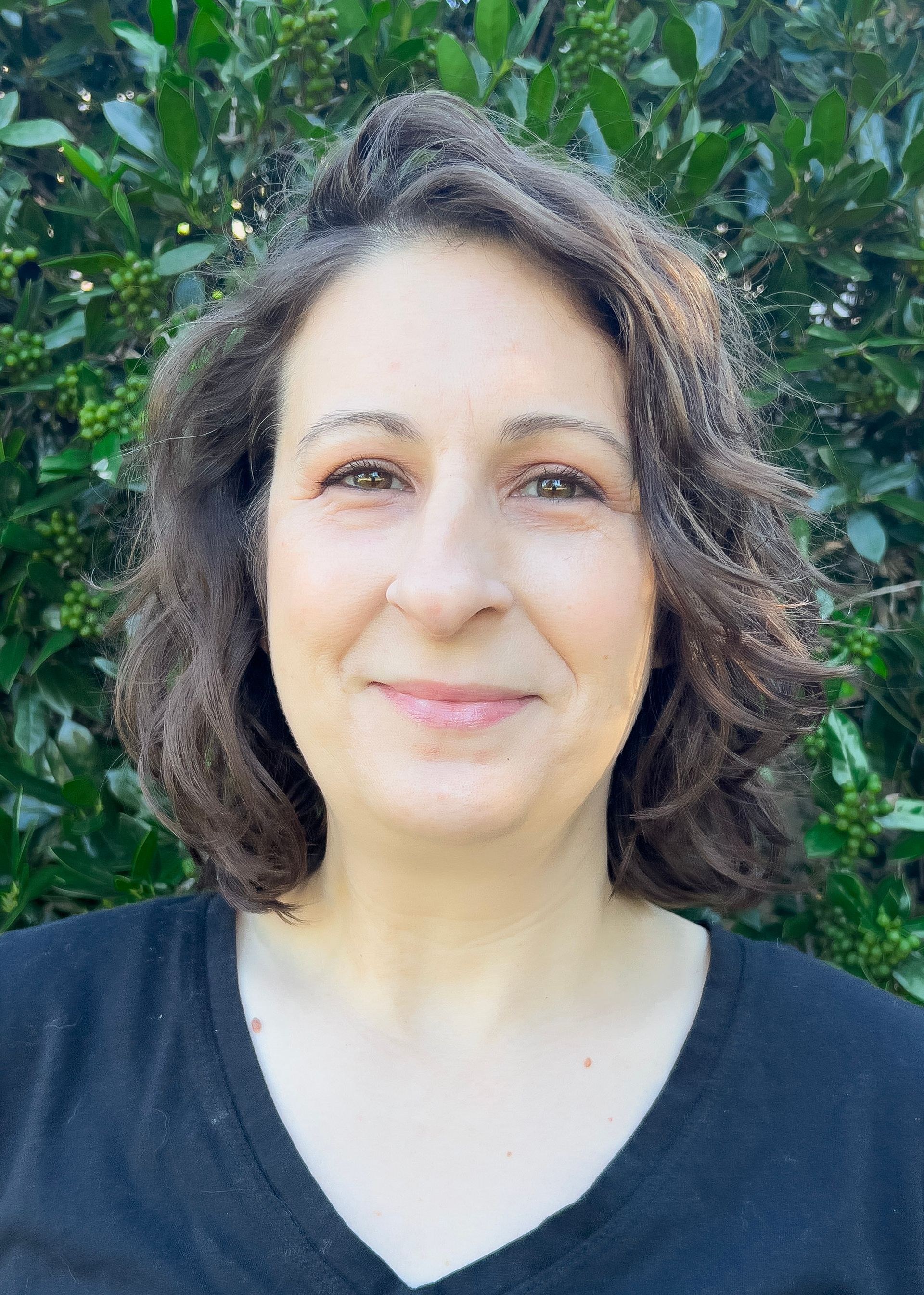 Woman with short brown hair smiling, wearing a black shirt, standing in front of greenery.